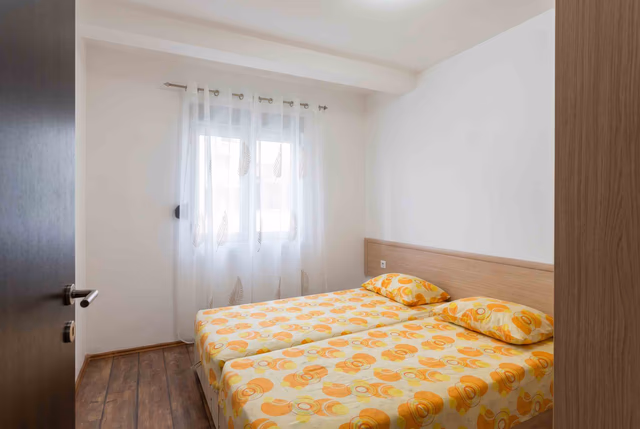 Gemütliches Schlafzimmer mit zwei Betten in einer Wohnung in Bečići, Montenegro: helle Einrichtung, Holzboden und Fenster mit Tüllgardine.