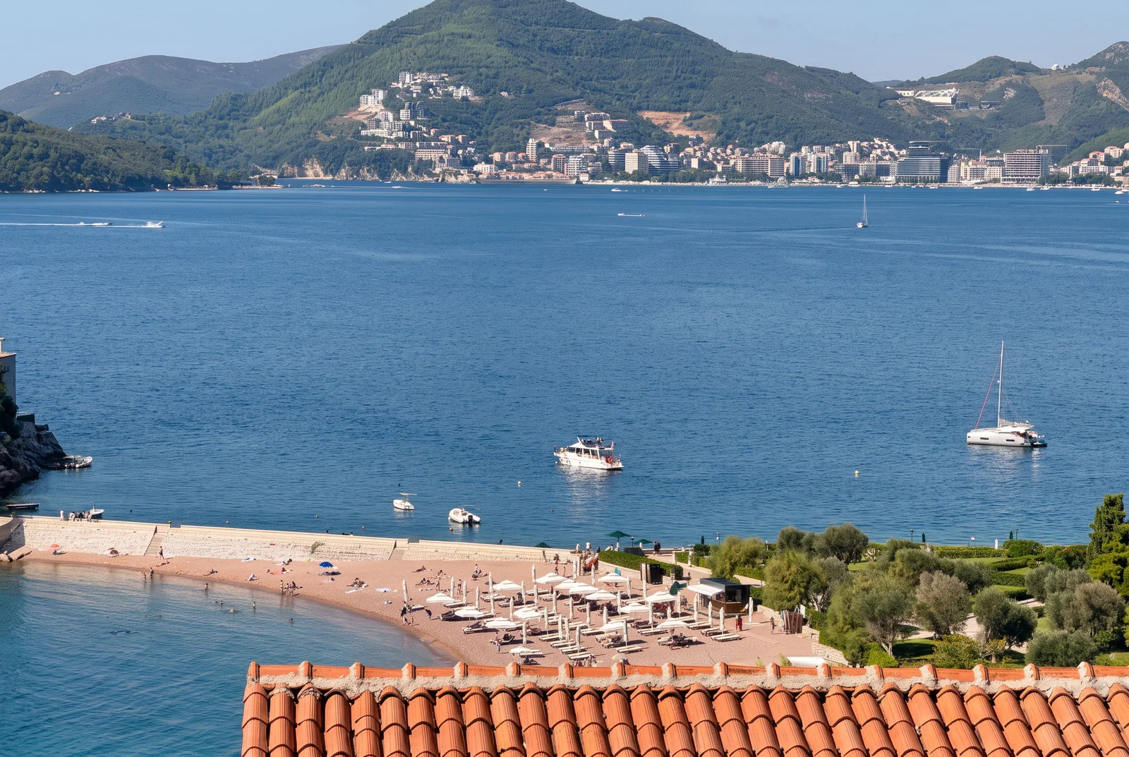 Pogled na more, plažu sa suncobranima, brodove i grad na brdu, terakotna crep u prvom planu.