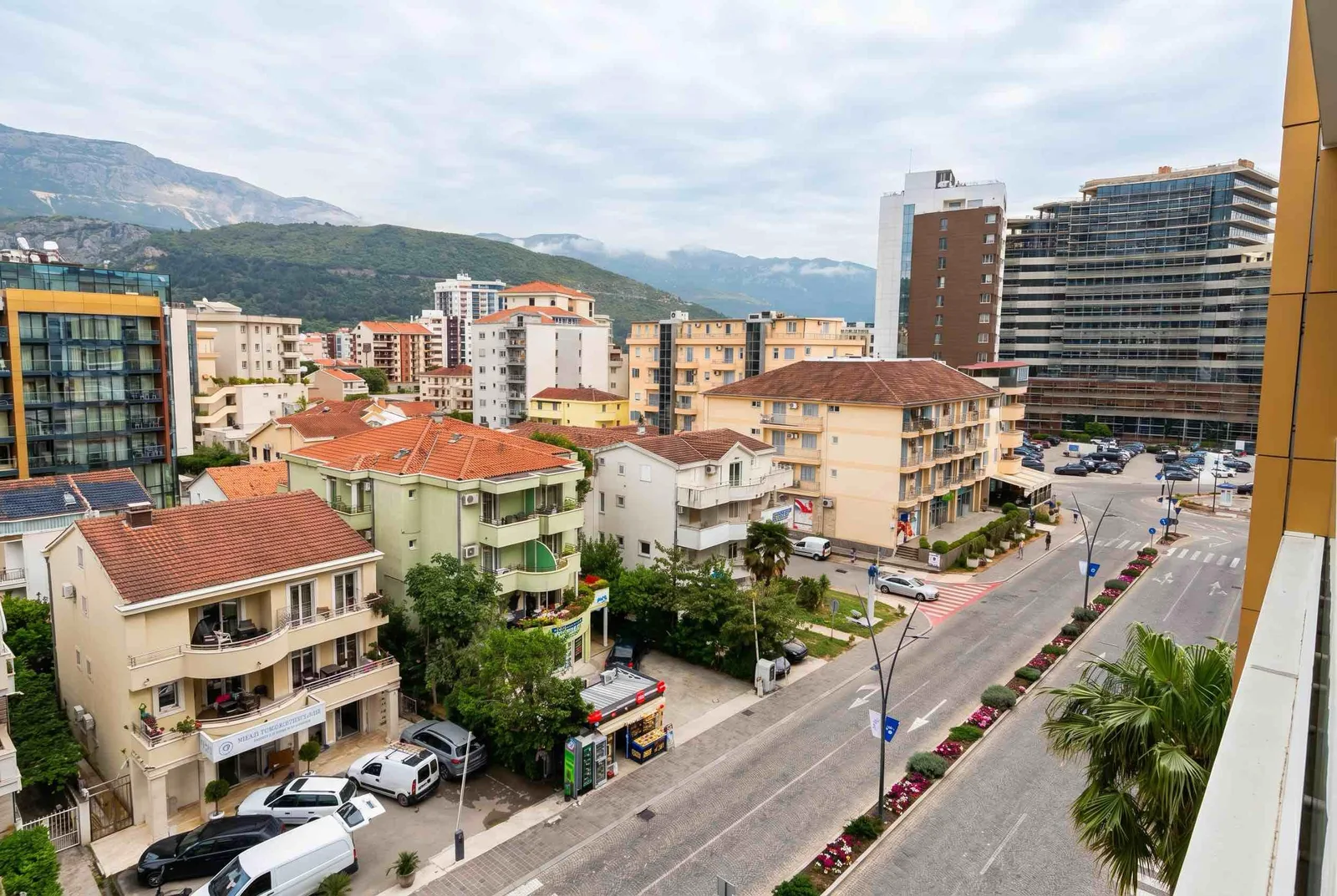 Panoramic view of a cityscape with multi-story buildings, a street, and mountains in the background.