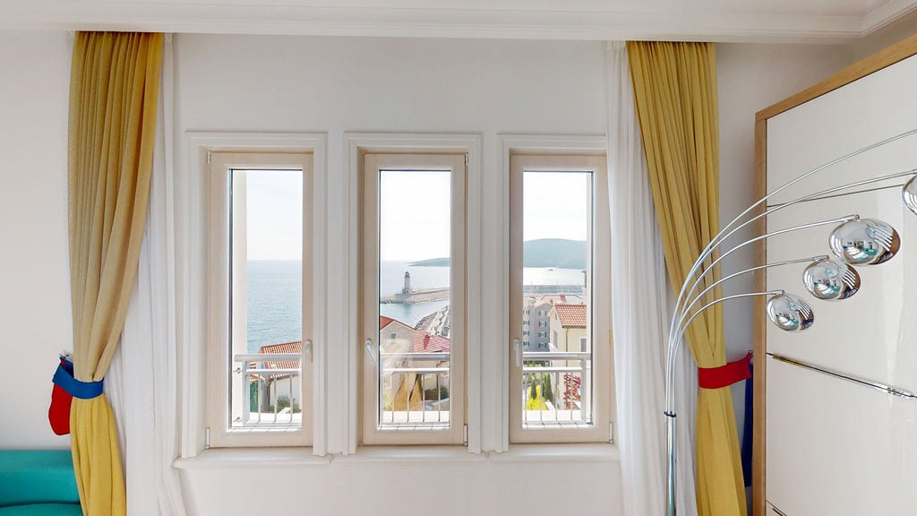 Karadağ, Luštica'daki dairenin oturma odası penceresinden deniz ve deniz feneri manzarası: sarı perdelerle aydınlık iç mekan.