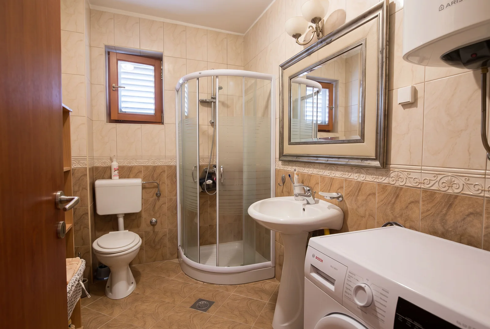 Salle de bain avec cabine de douche, toilette, lavabo et machine à laver sur carreaux beiges.