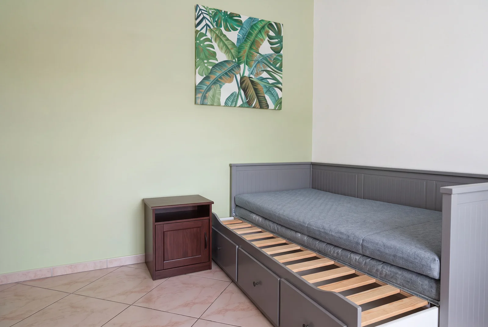 Grey sofa bed with a pulled-out sleeping frame and a dark wood nightstand against a light green wall with tropical art.