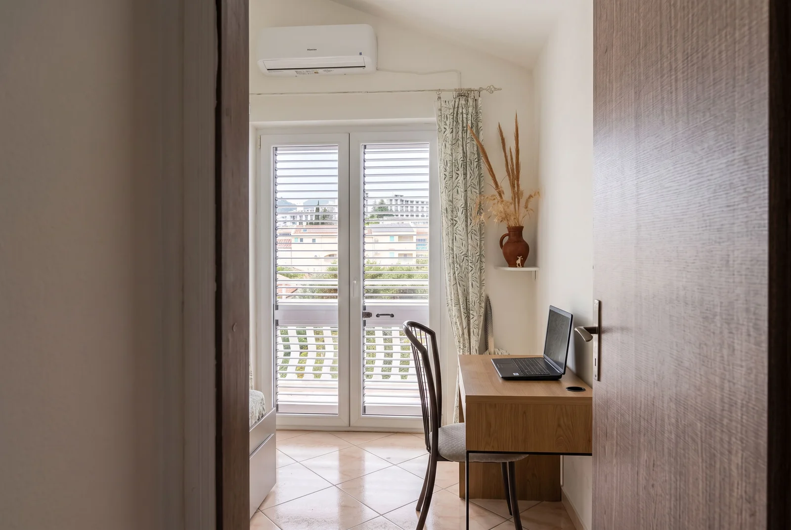 Part of a bedroom with a wooden desk, laptop, chair, and window with blinds visible.