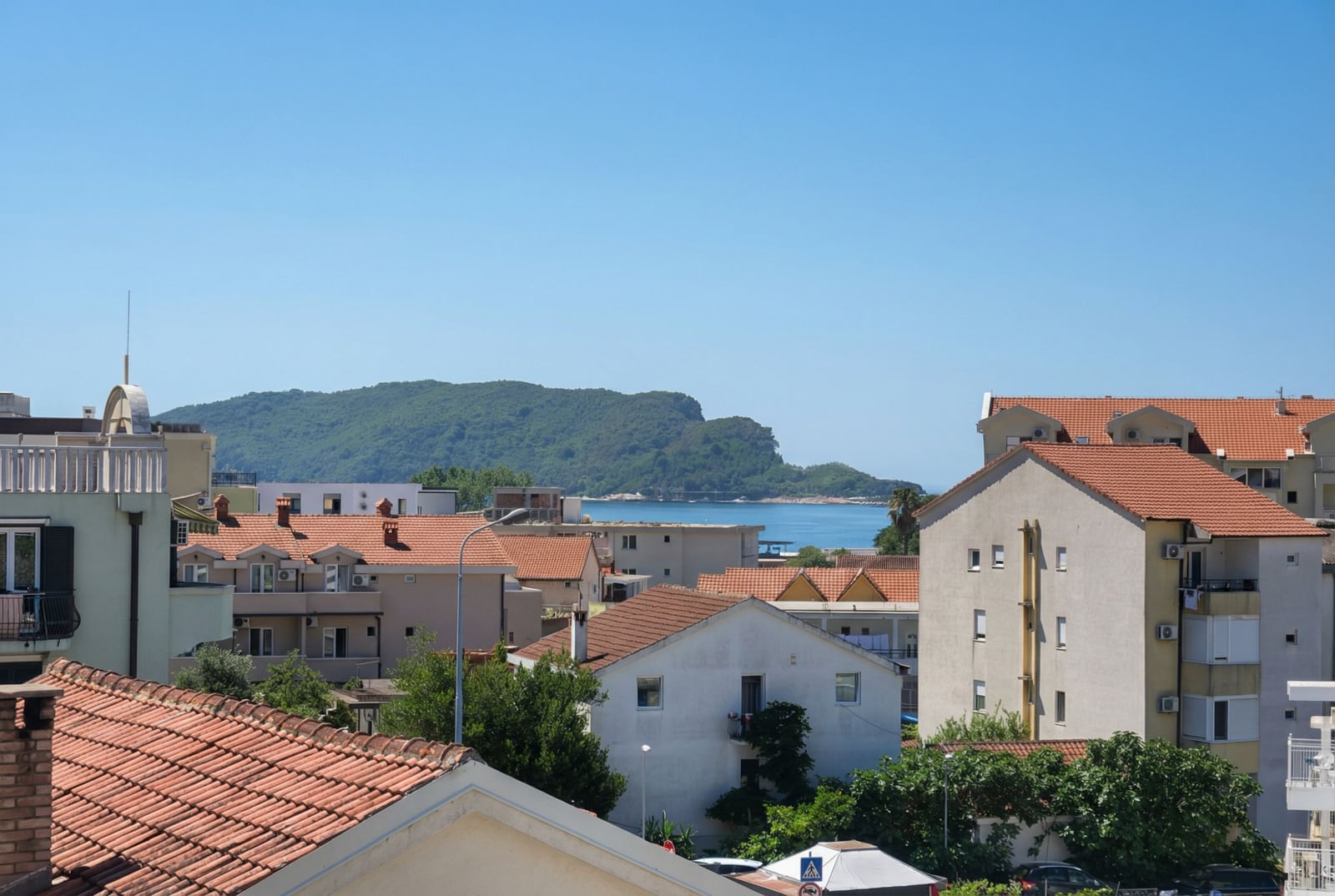 Pogled na more i planine iz prozora stana u Budvi, Crna Gora: panorama grada sa krovovima od crepa i zelenim brdima.