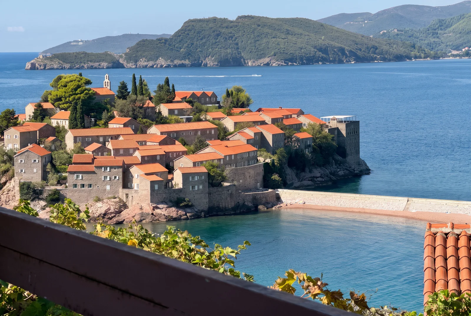 Pogled na kameni otok sa crvenim krovovima okružen morem i planinama.
