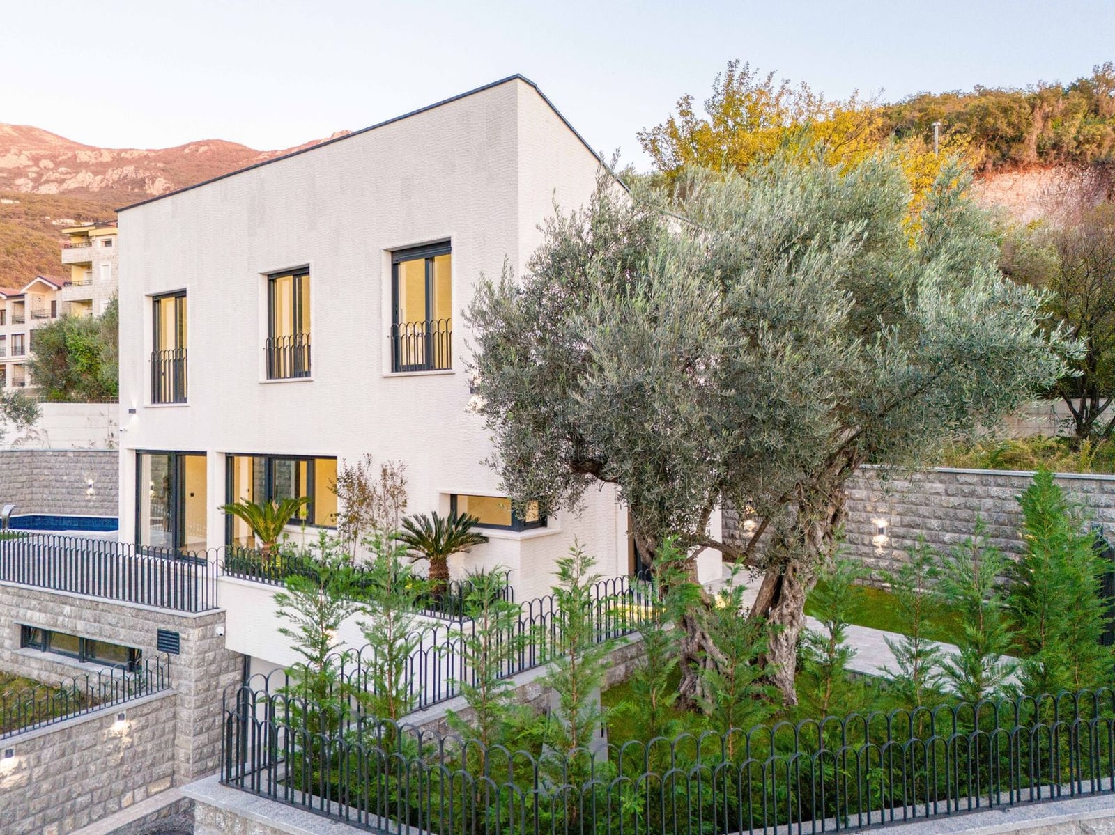 Villa blanche moderne à Kamenovo, Monténégro : façade avec fenêtres panoramiques, piscine, jardin avec olivier et clôture.