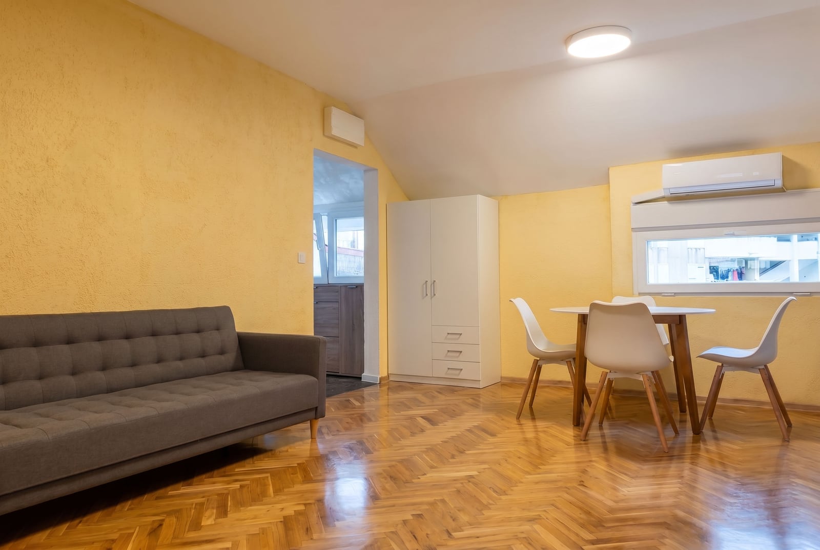 Living room in a Budva apartment, Montenegro: gray sofa, dining area with white chairs, parquet flooring, and yellow walls.
