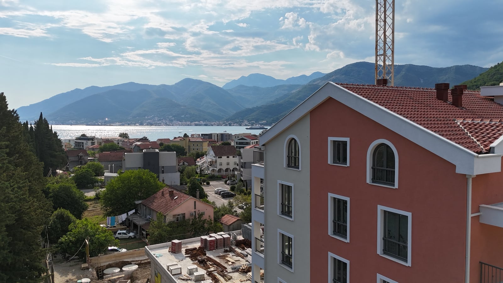 Pogled na more i planine iz prozora stana u Tivtu, Crna Gora: moderna stambena zgrada sa krovom od crepa i panorama grada.