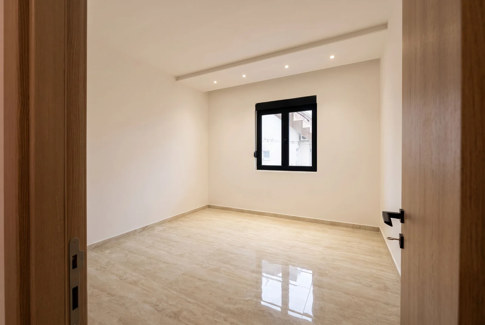 Empty room with beige tiles and a black window, viewed through a wooden doorway.