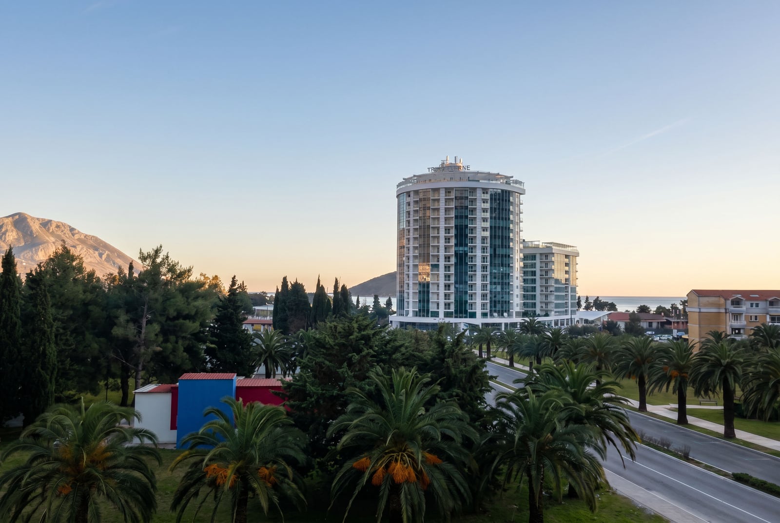 Modern Dukley Gardens residential complex in Budva, Montenegro, with a view of palm trees, mountains, and the coastline.