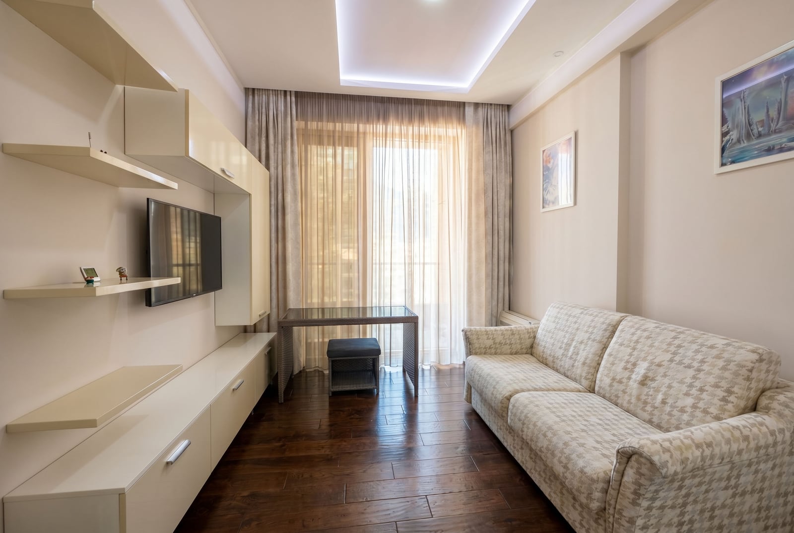 Modern living room in a Budva apartment, Montenegro: light-colored furniture, sofa, TV area, and panoramic window with balcony access.