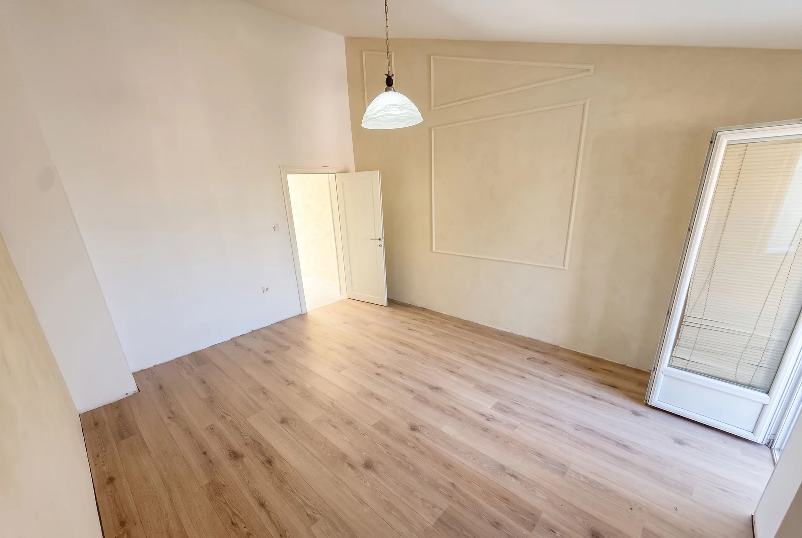 Pusta soba sa laminatom, visećom lampom i dekorativnim panelom na bež zidu.