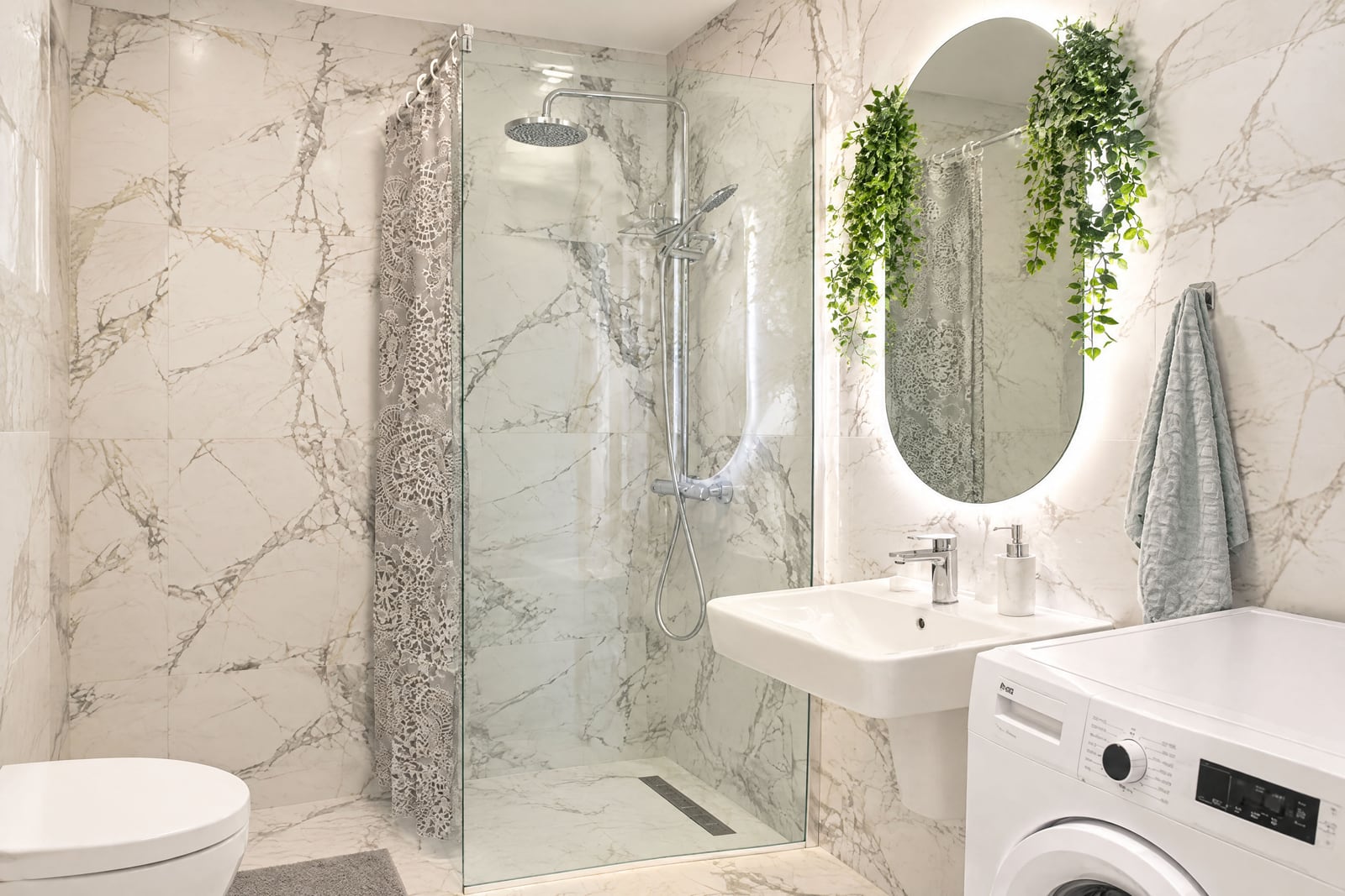 Bathroom with marble tiles, glass shower enclosure, sink, backlit mirror, and washing machine.