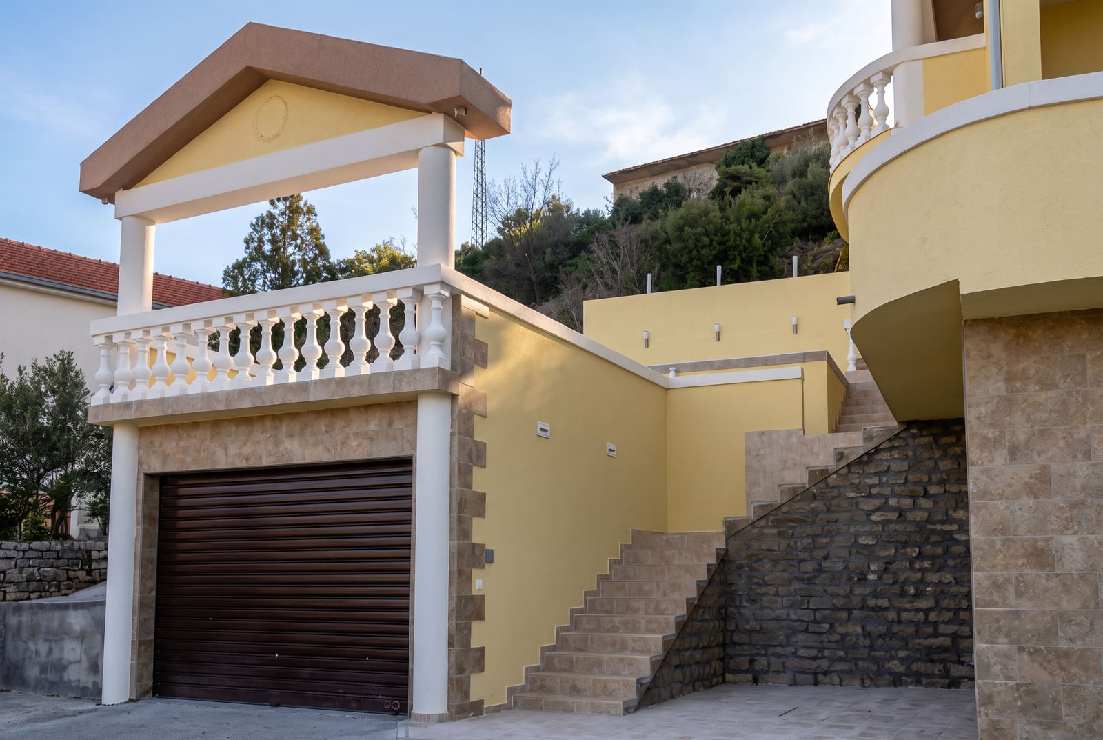 House facade in Tivat, Montenegro: garage with roller shutters, terrace with balustrade, and stone staircase