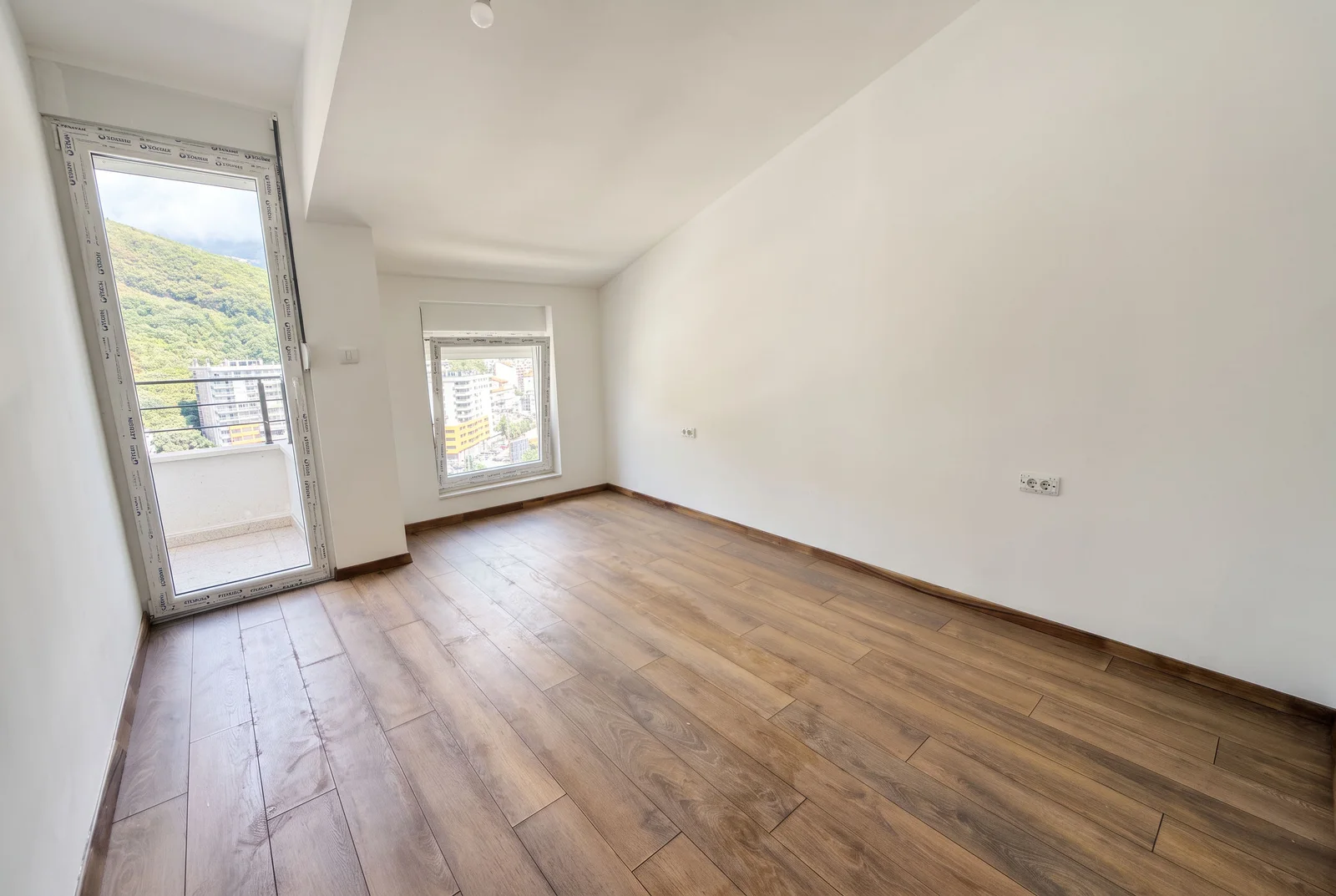 Pusta soba sa drvenim podom, belim zidovima, prozorom i vratima na balkon sa pogledom na brda.