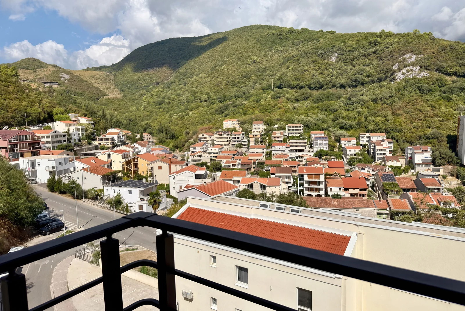 Prikaz s balkona na kuće sa crvenim krovovima i zeleni šumoviti brdski pozadina.