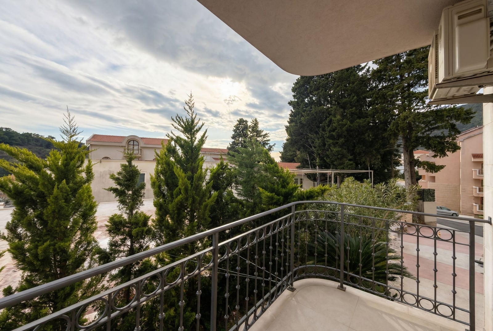 View from the apartment balcony in Petrovac, Montenegro: wrought-iron railings, green coniferous trees, and a panoramic view of the residential complex.