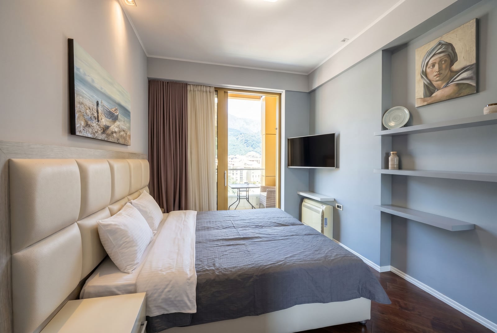 Modern bedroom in a Budva apartment, Montenegro: double bed, TV, balcony with mountain views, and stylish decor.
