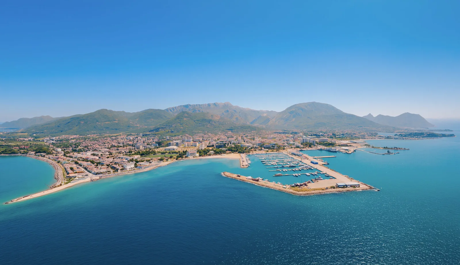 Yat limanı, plaj ve arka planda dağlar olan bir kıyı şehrinin hava fotoğrafı.