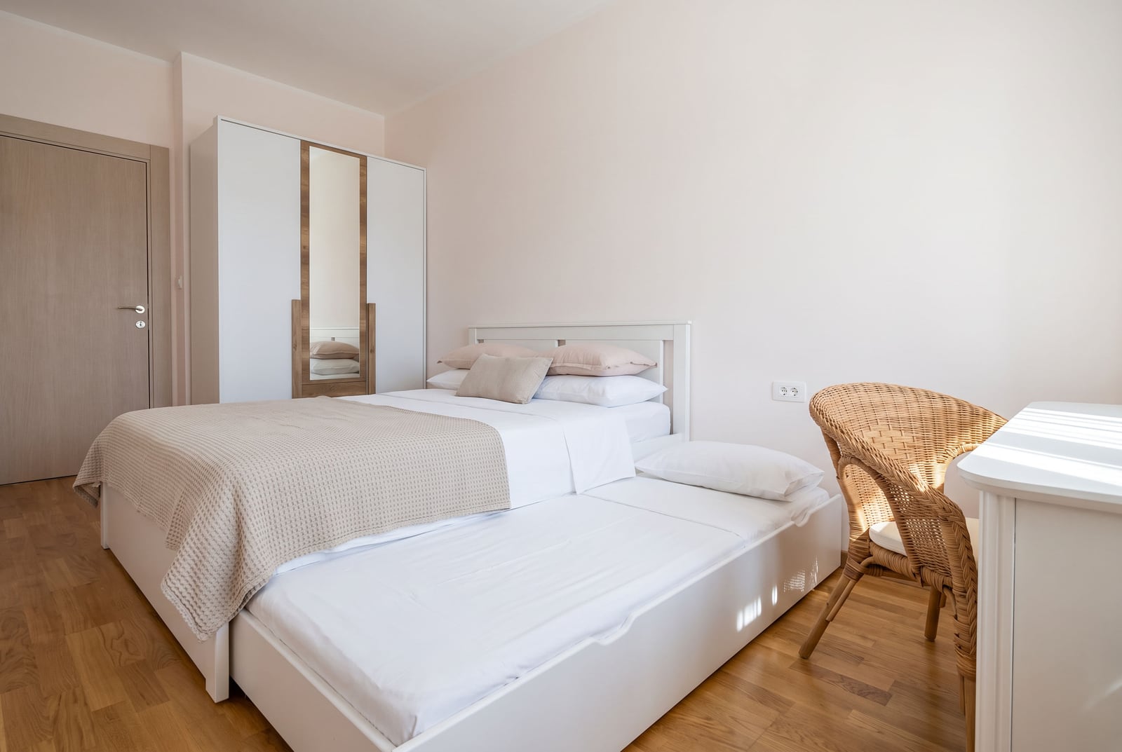 Светлая спальня в квартире в Будве, Черногория: двуспальная кровать с выдвижным местом, шкаф с зеркалом и плетеный стул.