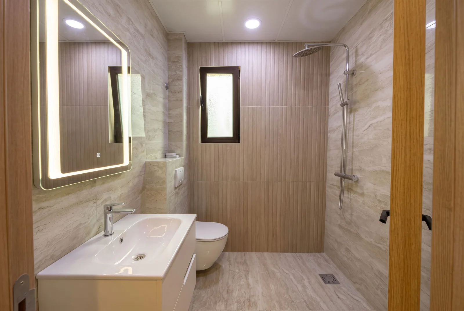 Modern bathroom featuring a sink, wall-mounted toilet, shower system, and an illuminated vanity mirror.