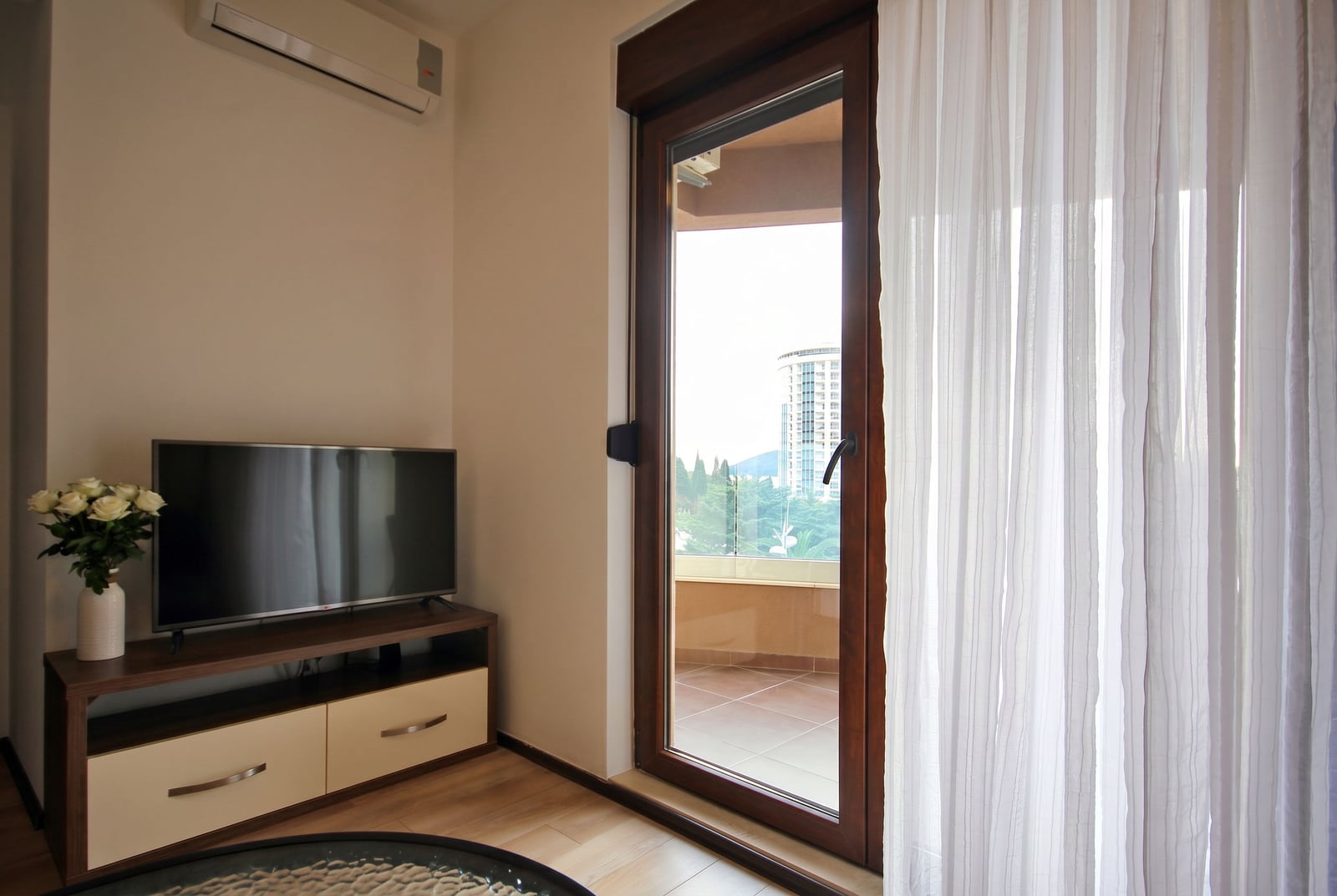 Living room in a Budva apartment, Montenegro: TV area, modern furniture, and access to a balcony with a panoramic city view.