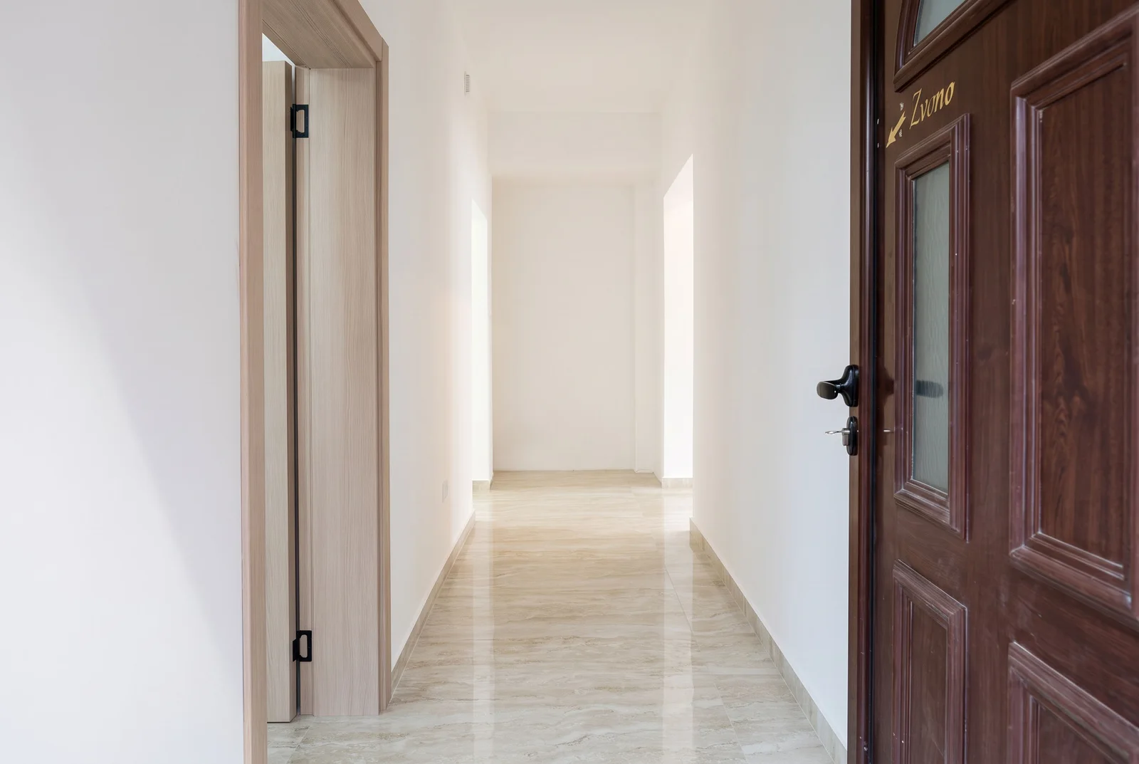 A long, bright hallway with polished stone flooring, white walls, and a dark wooden entrance door on the right side.