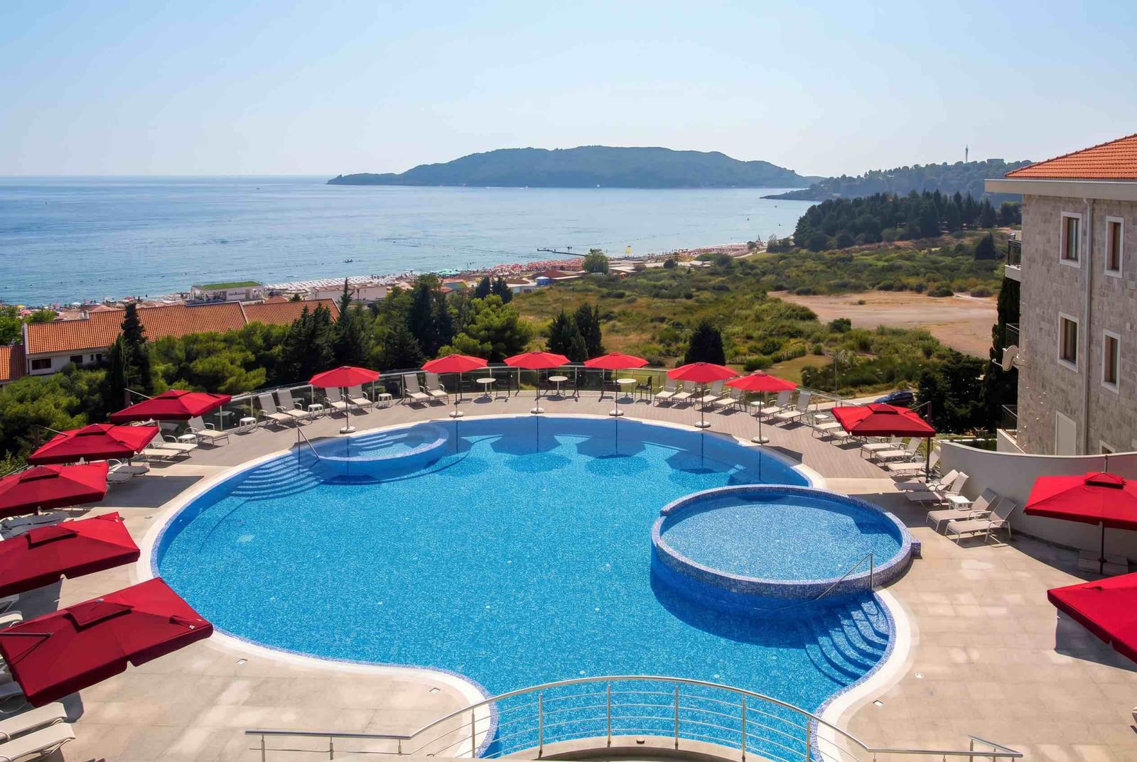 Outdoor pool with red umbrellas and sea view in Bečići, Montenegro: a lounge area with sunbeds by the residential complex.