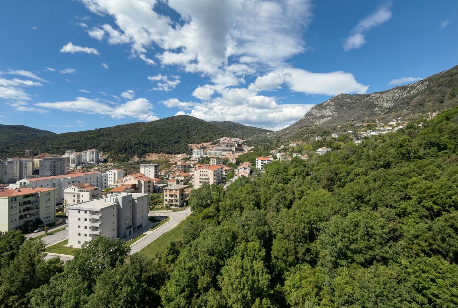 Panoramski pogled na stambeni deo Budve, Crna Gora: moderne zgrade, zeleni brežuljci i planine pod vedrim plavim nebom.