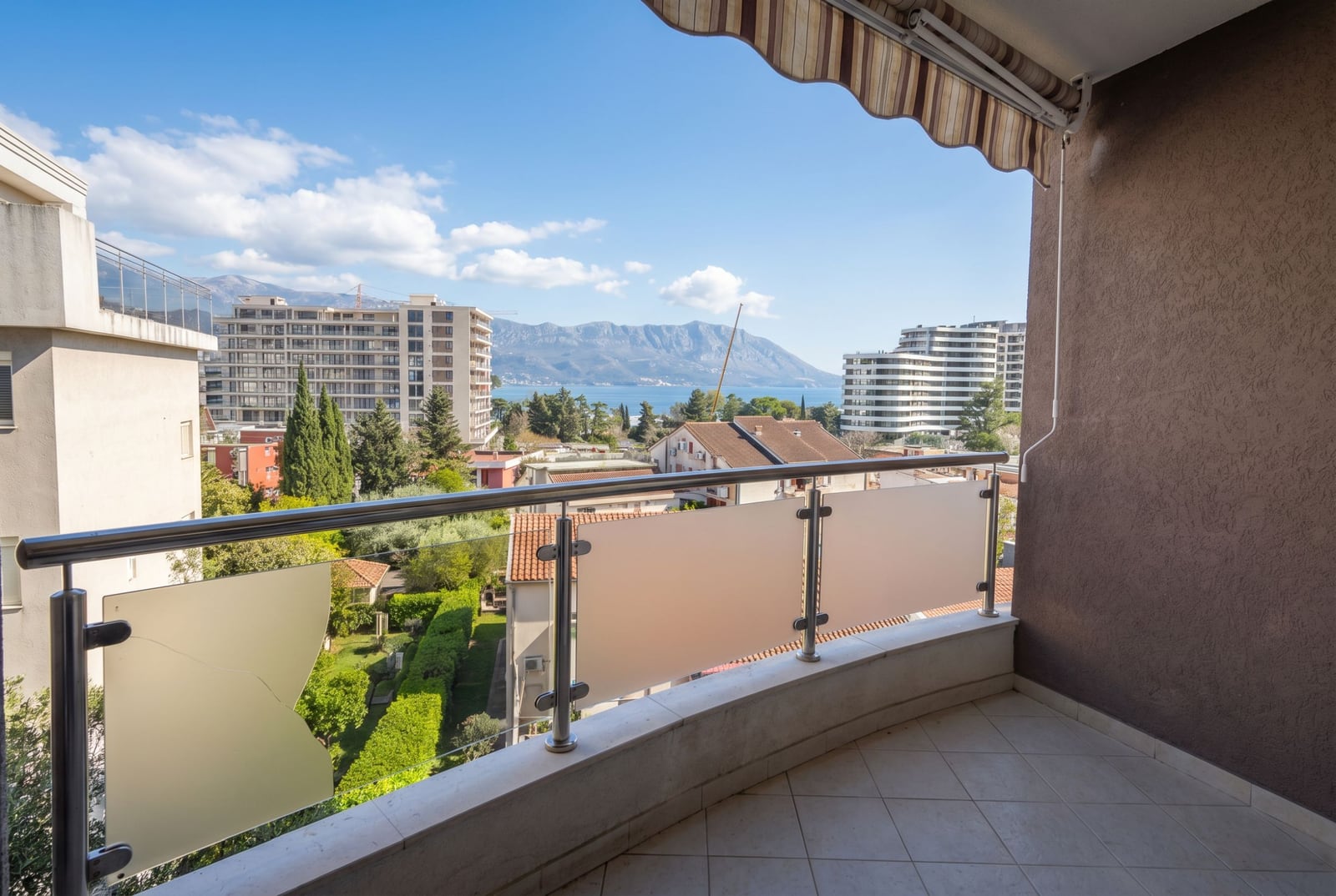 Balkon stana u Budvi, Crna Gora, sa panoramskim pogledom na more, planine i grad, zaštitnim nadstrešnicom i staklenom ogradom.