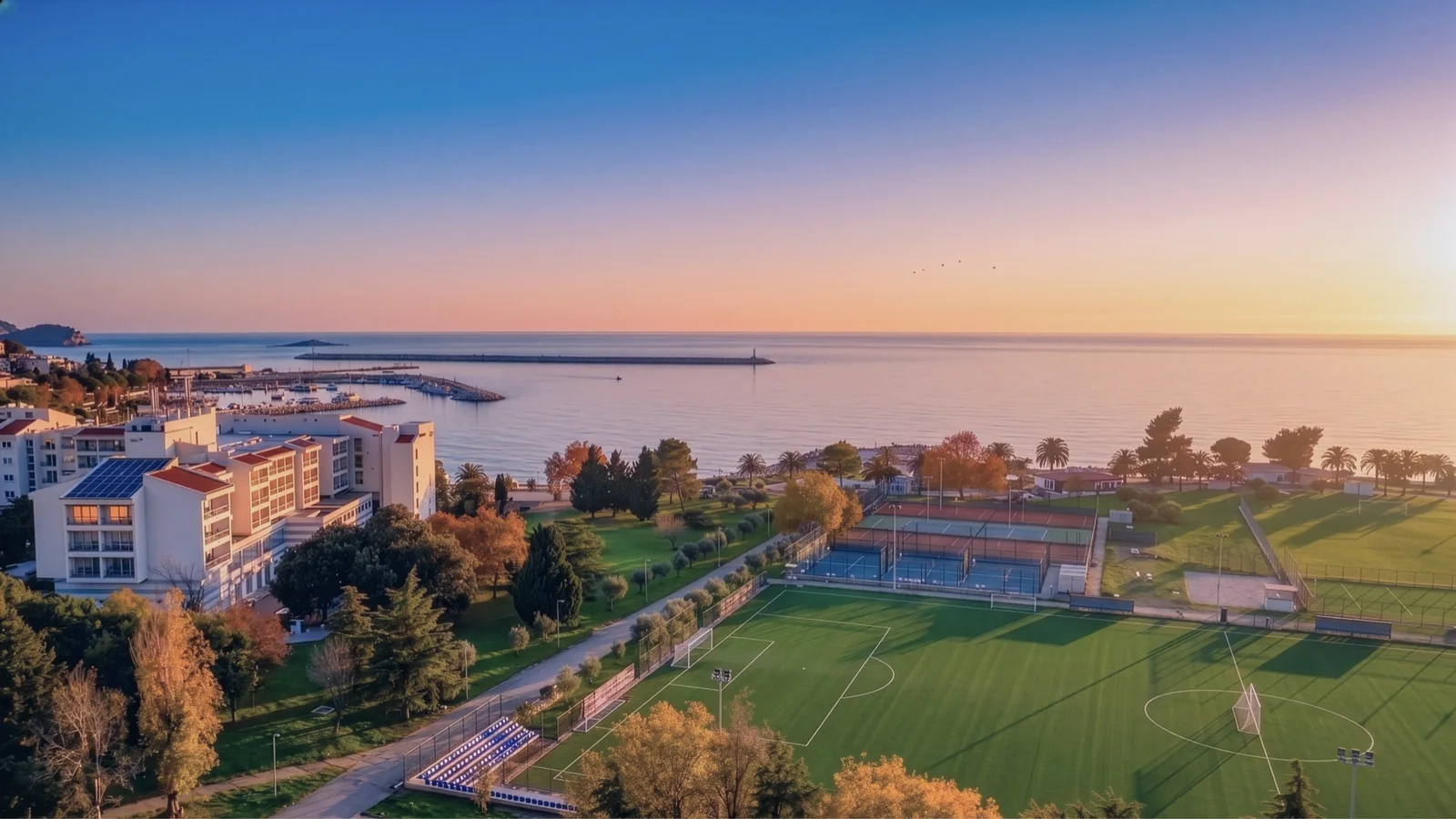 Güneş batarken denize yakın bir futbol sahası ve tenis kortları olan spor kompleksinin havadan görünümü.