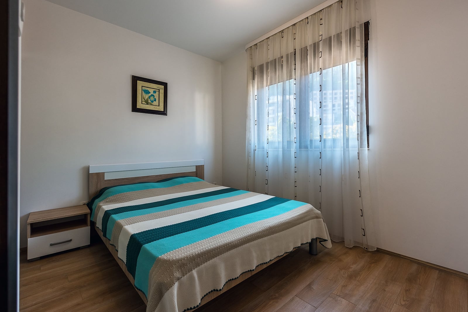 Bright bedroom with a double bed in an apartment in Budva, Montenegro: laminate flooring, bedside table, and a window with sheer curtains.