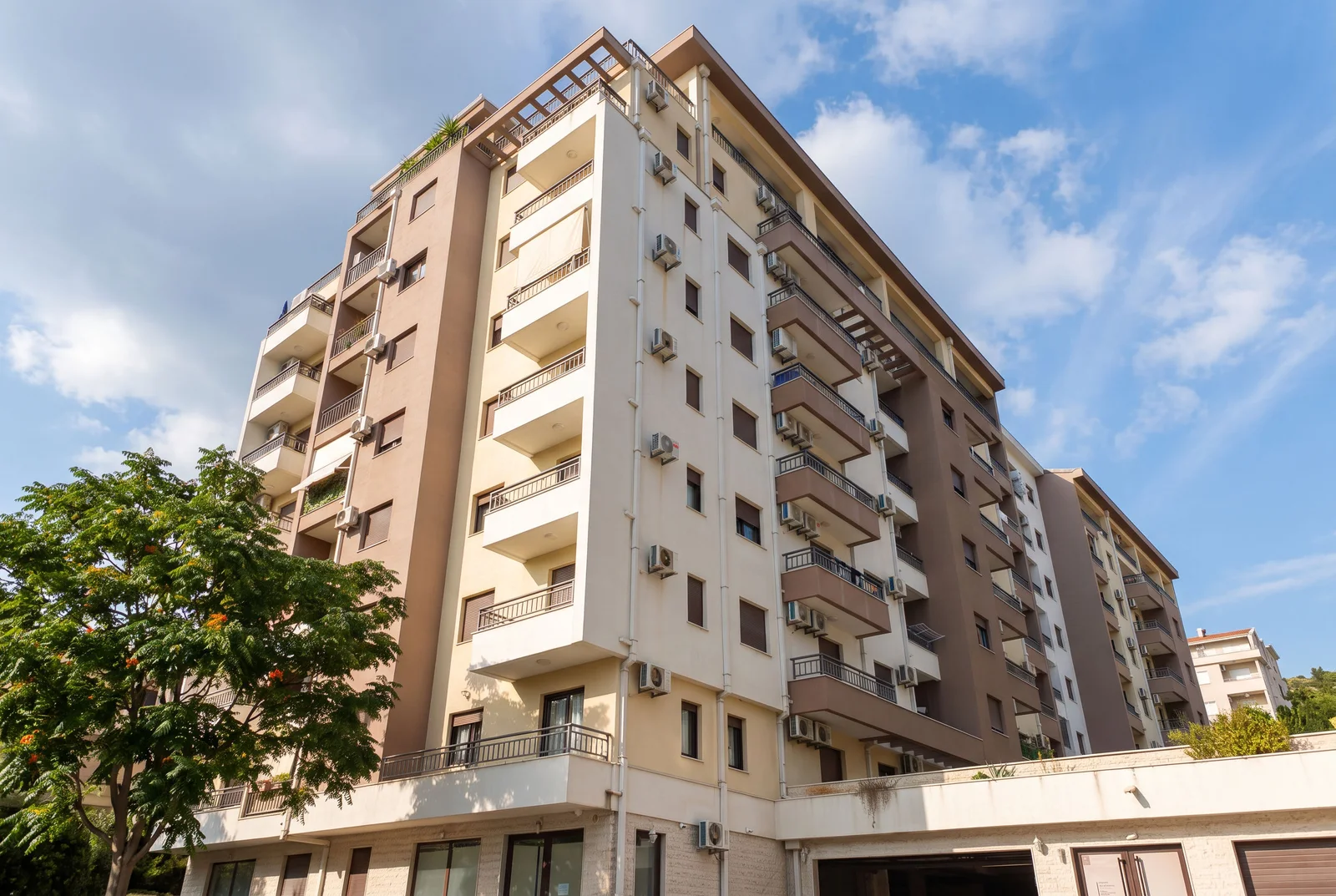 Exterior view of a multi-story residential building featuring balconies and AC units against a blue sky.