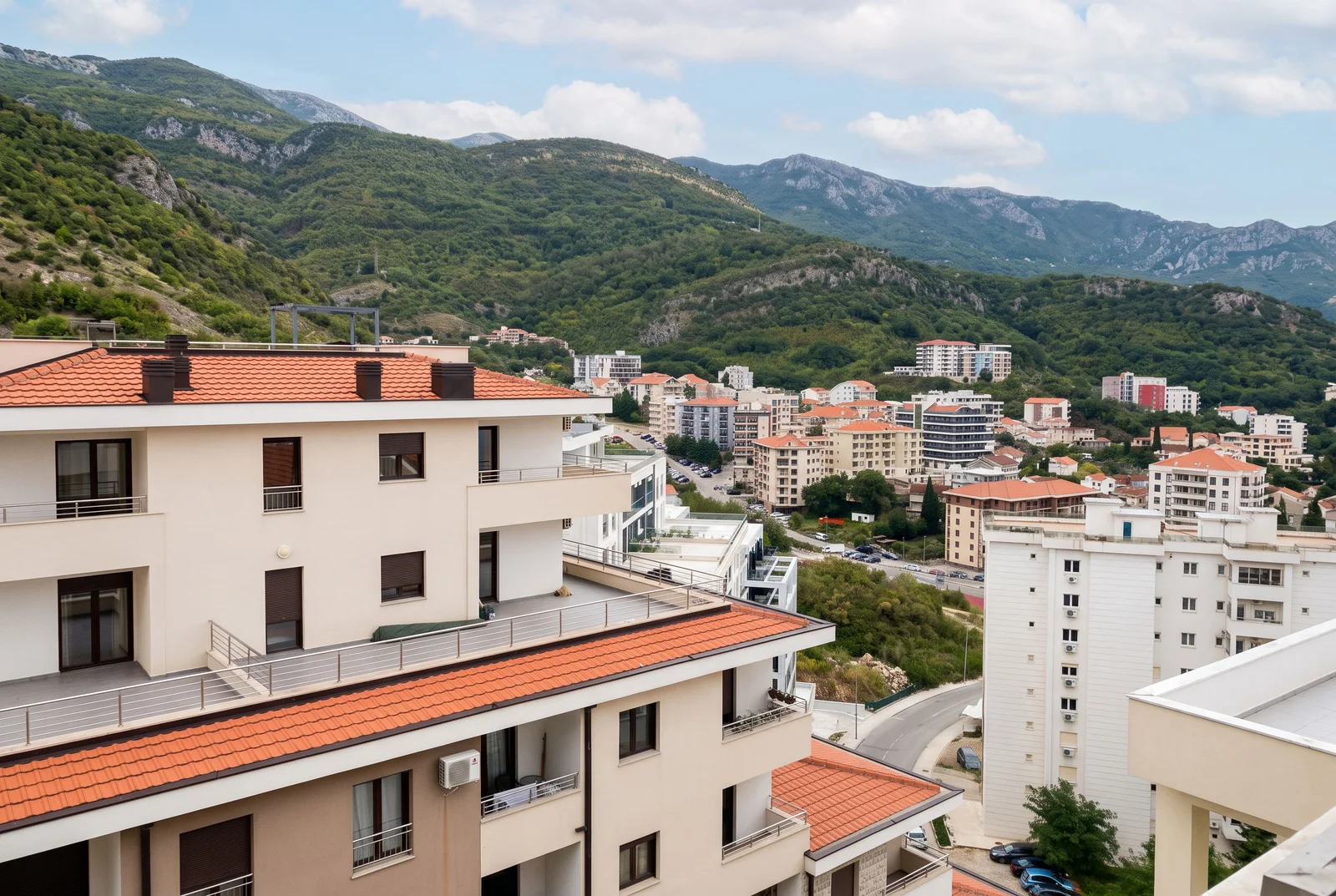 Prikaz gradskih zgrada sa crvenim crepanim krovovima i terasama na pozadini zelenih planina.