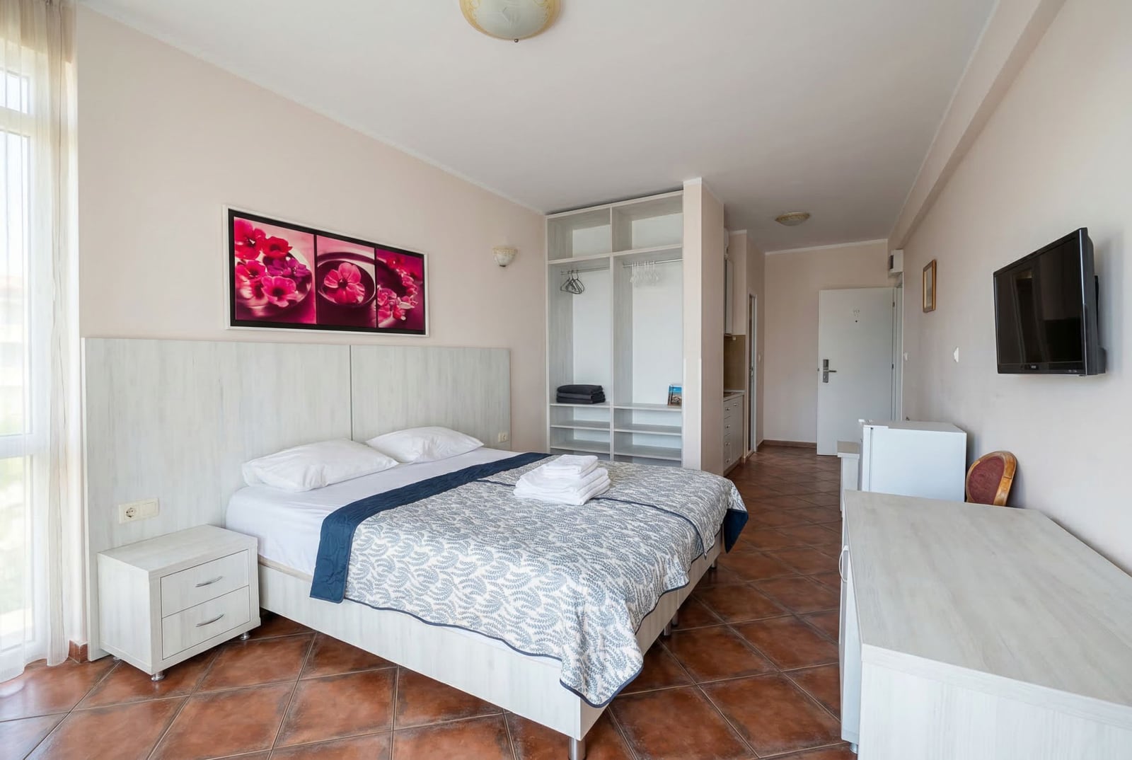 Svetla spavaća soba u hotelu Bečići, Crna Gora: bračni krevet, otvoreni ormar, TV i pločice na podu u sobi.