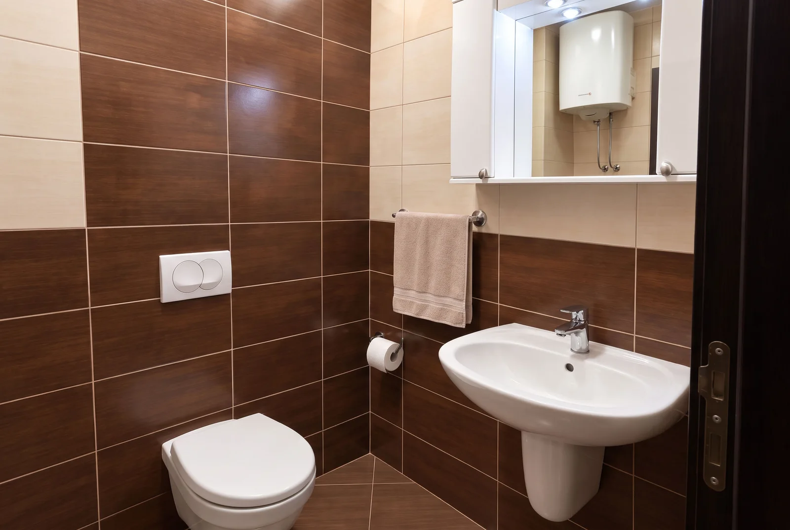 Bathroom featuring a white toilet, wall-mounted sink, mirrored cabinet, and beige towel against brown tiles.