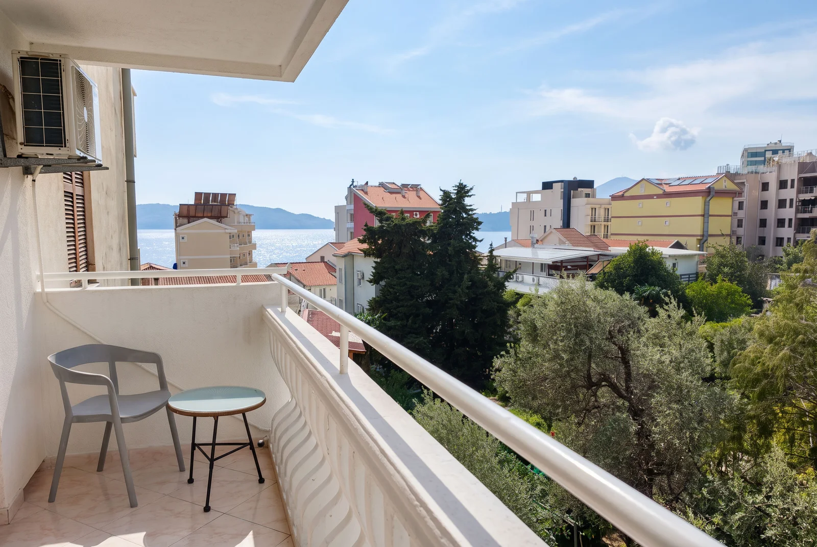 Balcony with grey plastic chair and small table overlooking sea and city buildings under blue sky.