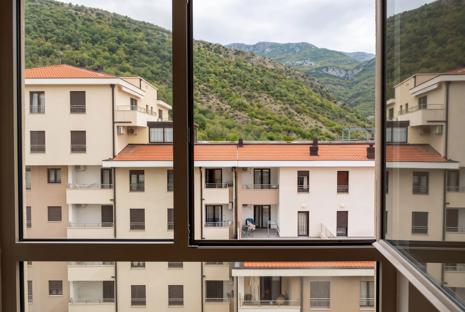 Prikaz kroz prozor na stambene zgrade s crvenim krovovima i zelenim planinama.