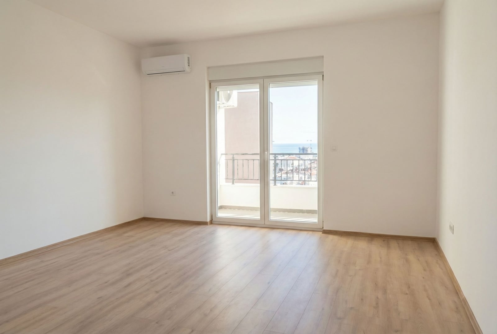 Svetla prazna dnevna soba sa laminatom, klima-uređajem i izlazom na balkon s pogledom na more u Budvi, Crna Gora.