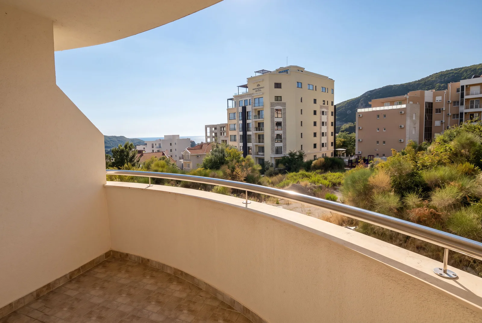 Prazan balkon sa pločanim podom i metalnom ogradom sa pogledom na hotel i brda.