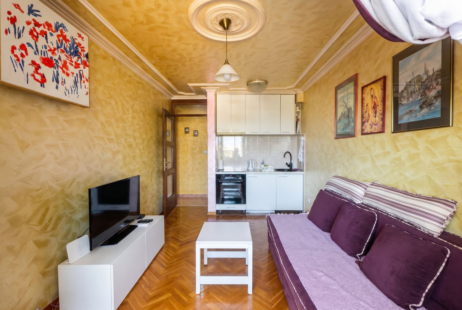 Living room with kitchen in an apartment in Budva, Montenegro: purple sofa, white furniture, parquet flooring, and decorative wall finishes.