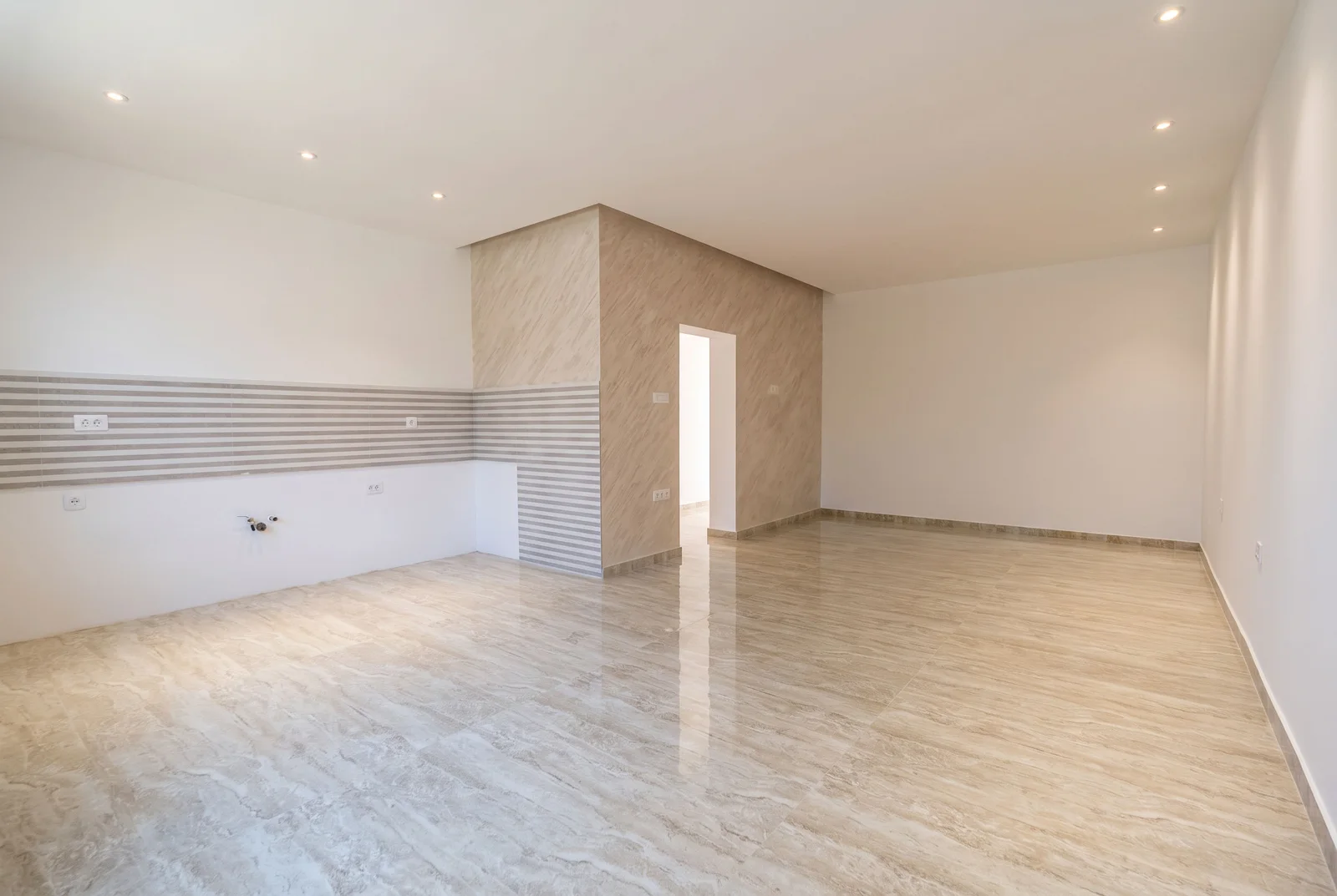 Empty room with glossy tiled floor and striped wall tiles, visible electrical outlets and pipe stubs, likely a kitchen under renovation.