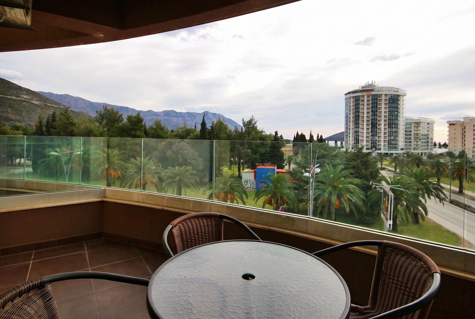 View from a balcony of an apartment in Budva, Montenegro: wicker furniture, glass railing, mountains and palm trees against the backdrop of the city.