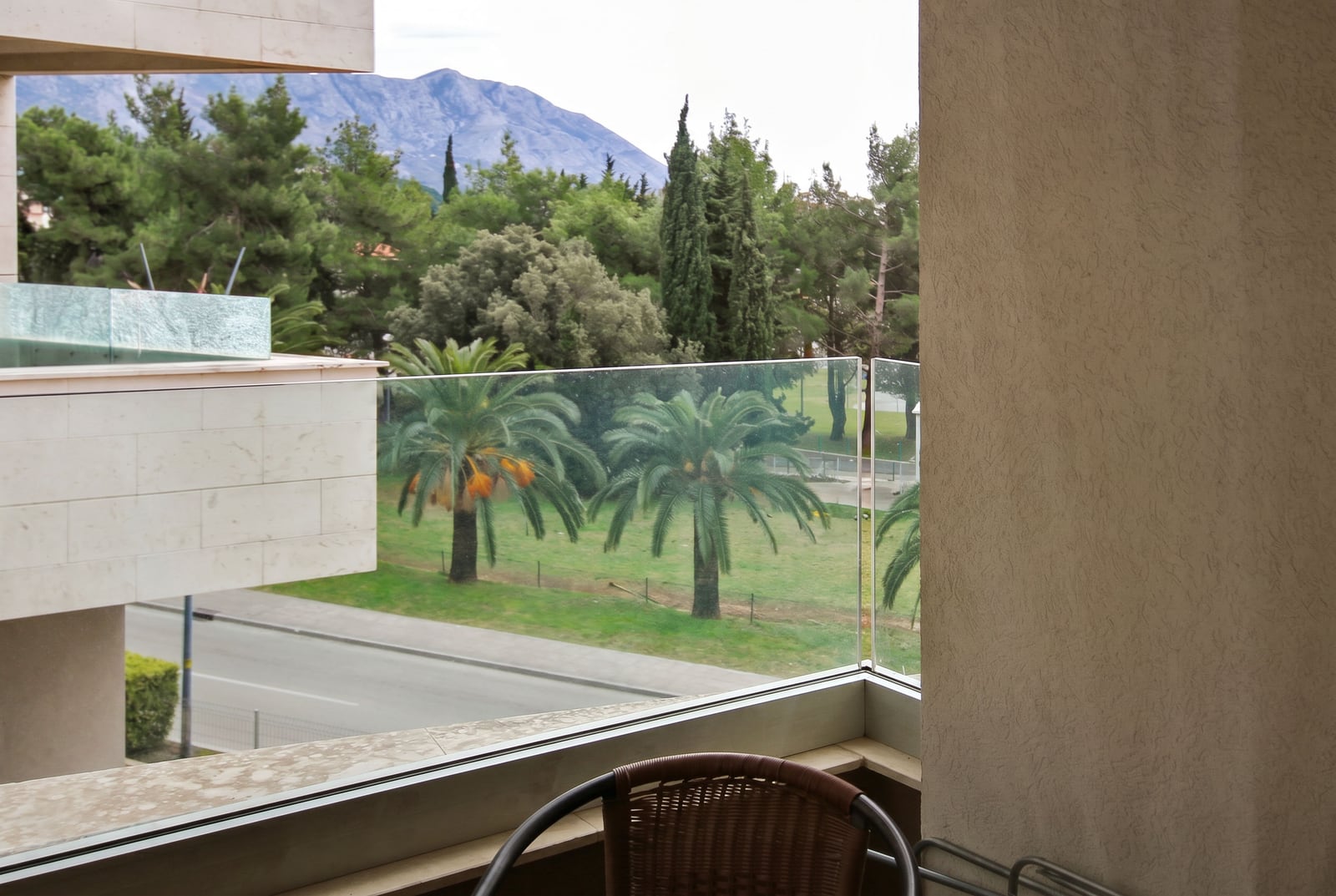 View from the apartment balcony in Budva, Montenegro: glass railing, wicker chair, palm trees, and mountains in the background.