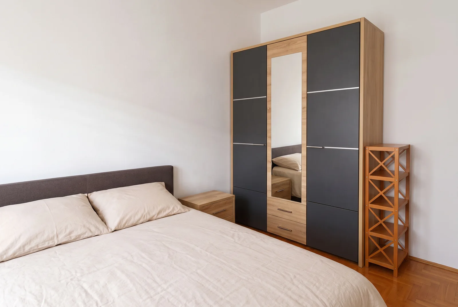 Bedroom featuring a double bed with beige linens, a grey and wood wardrobe with a mirror, and a wooden shelf unit on parquet flooring.