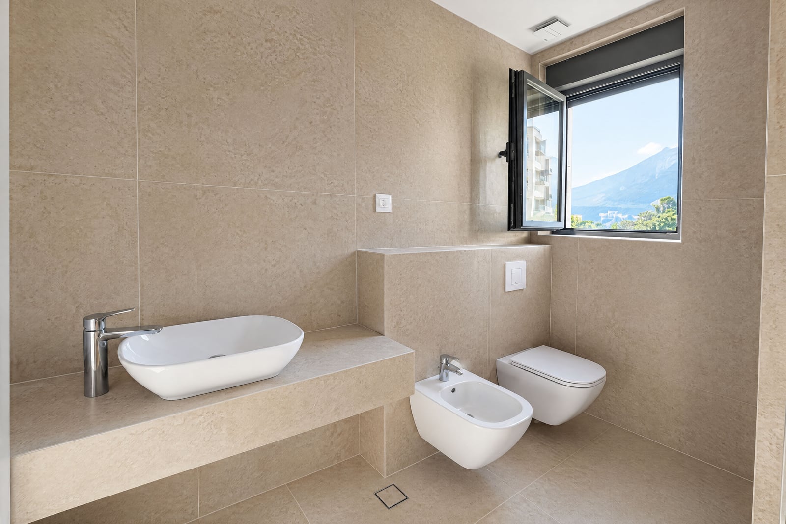 Salle de bain moderne avec sanitaires blancs, carrelage beige et fenêtre avec vue sur les montagnes dans un appartement, Bečići, Monténégro