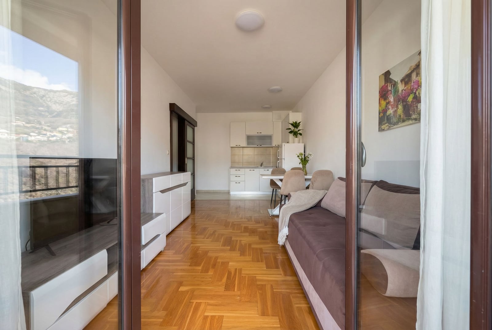 Dnevna soba sa kuhinjom u stanu u Budvi, Crna Gora: parket, moderan nameštaj, sofa i pogled na planine sa balkona.