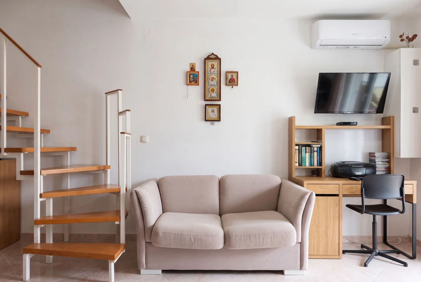 Living room featuring a beige sofa, wooden staircase, desk with TV, and religious icons on the wall.