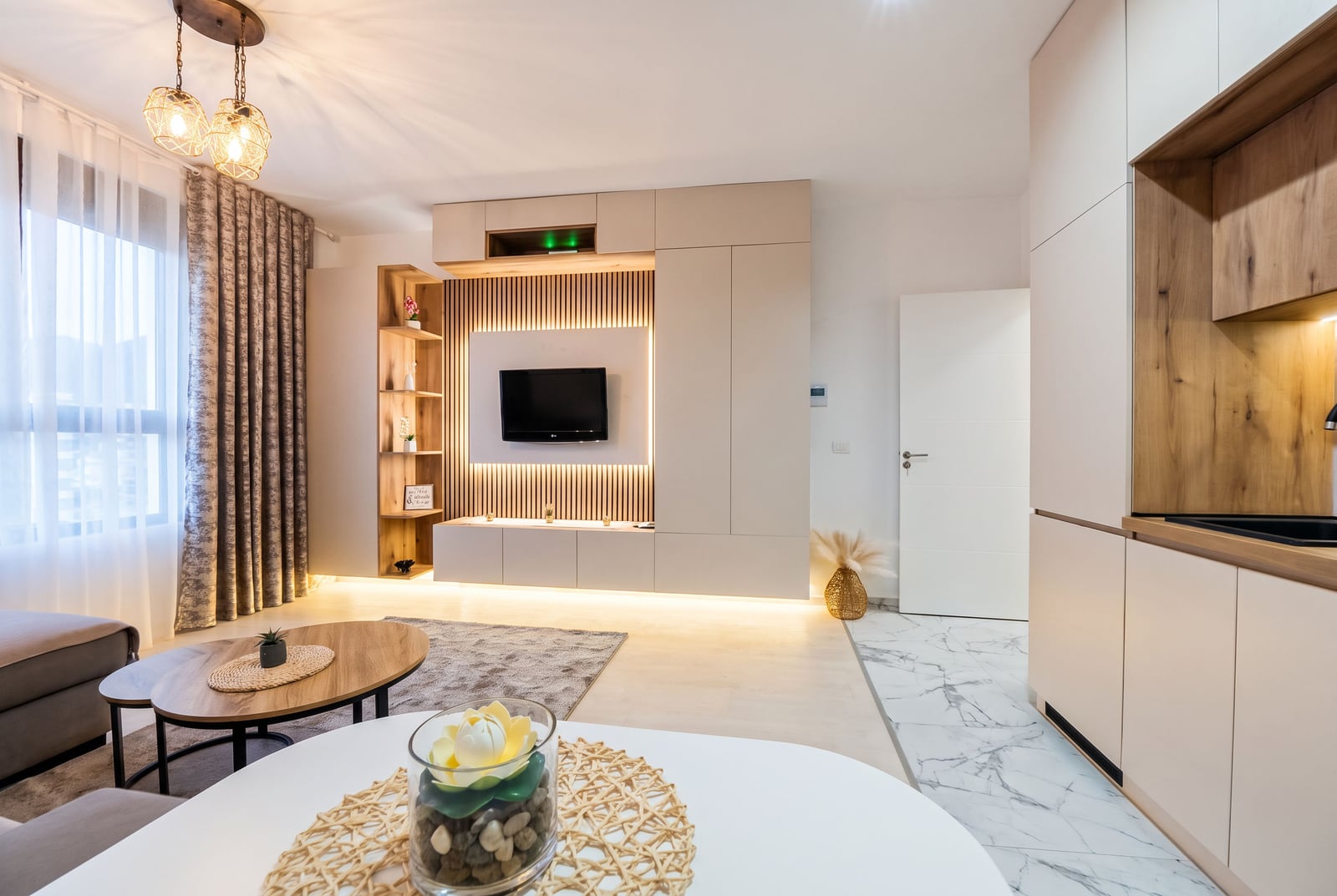 Modern living room featuring a TV on a slatted wood wall, sofa, coffee table, and an adjacent kitchen area visible on the right.