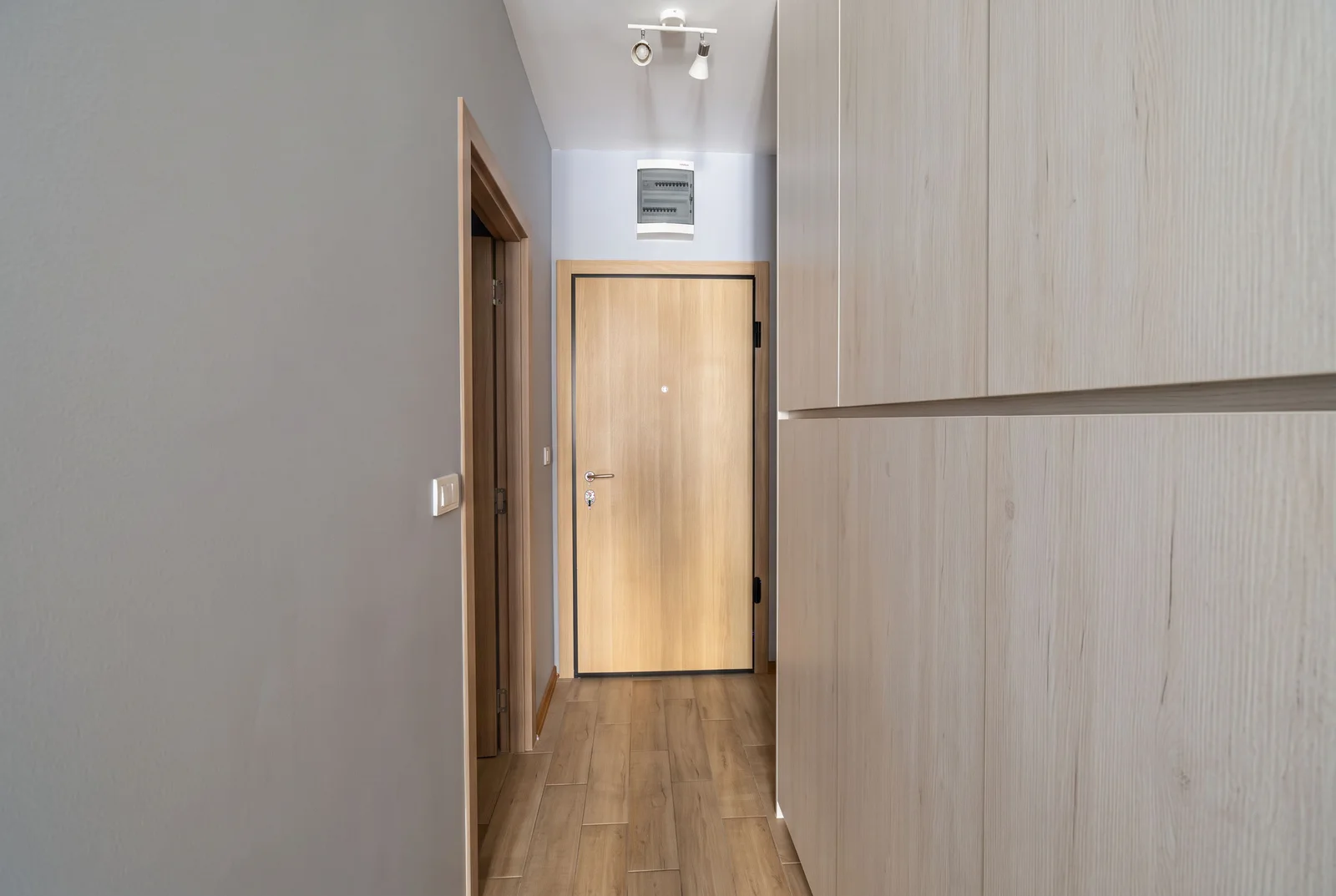 A narrow hallway featuring a wooden door, light wood cabinetry, and an electrical panel on the wall.